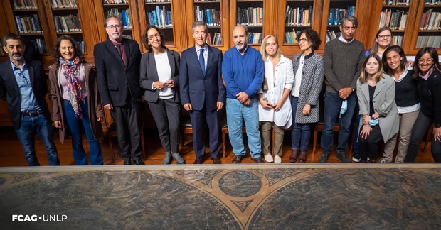 En la imagen se ve, en primer plano, el mapa del cielo sobre la extensa mesa de la BIblioteca de FCAG. En el fondo, están autoridades de FCAG-UNLP y de la UNSAM, así como miembros de esta Facultad y parte del grupo de profesionales involucrados en la restauración del mapa.