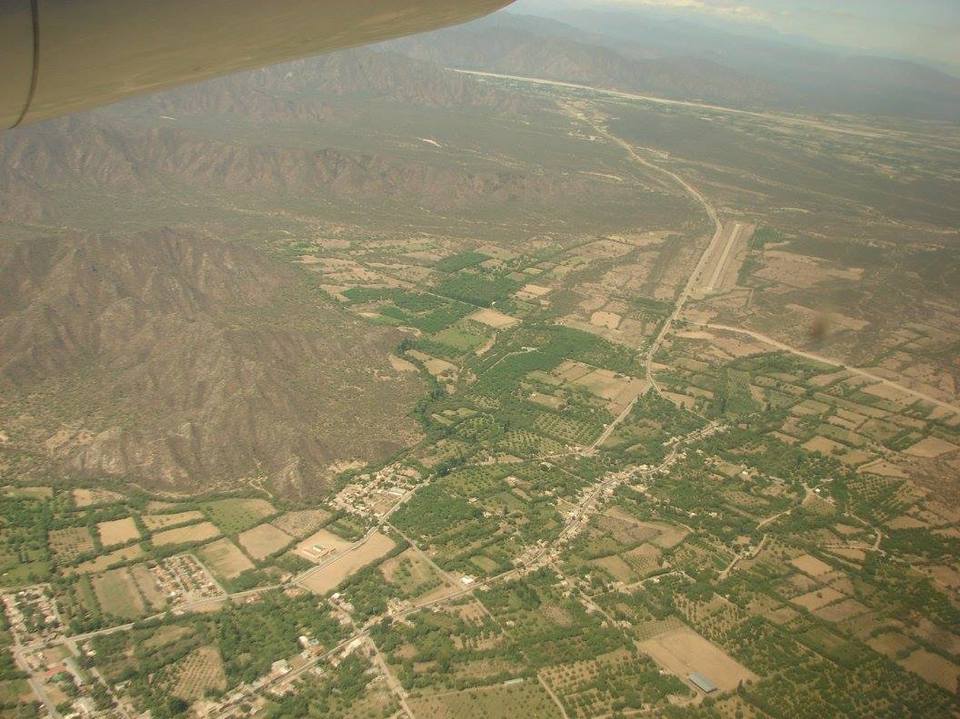 La imagen corresponde a un avista aérea del sitio arqueológico El Shincal. Se observan los restos de las edificaciones, cordones montañosos y parcelas verdes.